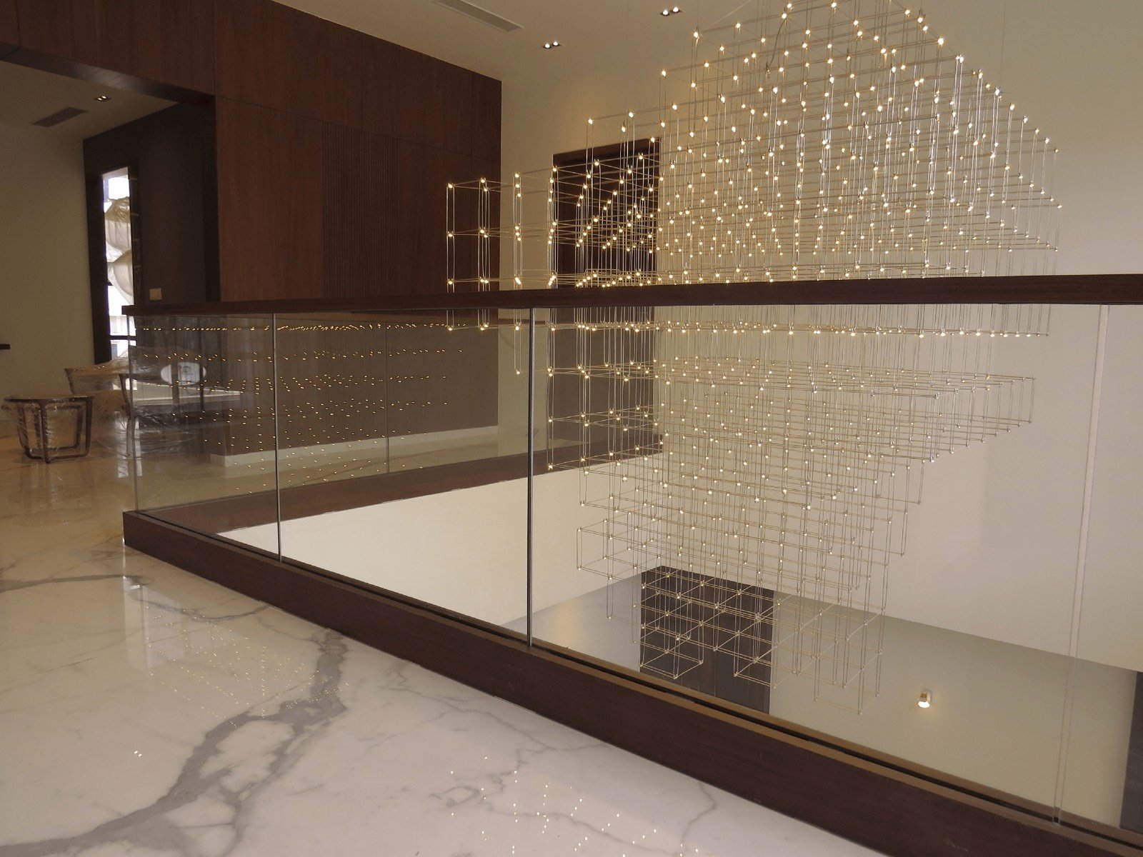 A frameless glass handrail along a marble mezzanine under a hanging light installation
